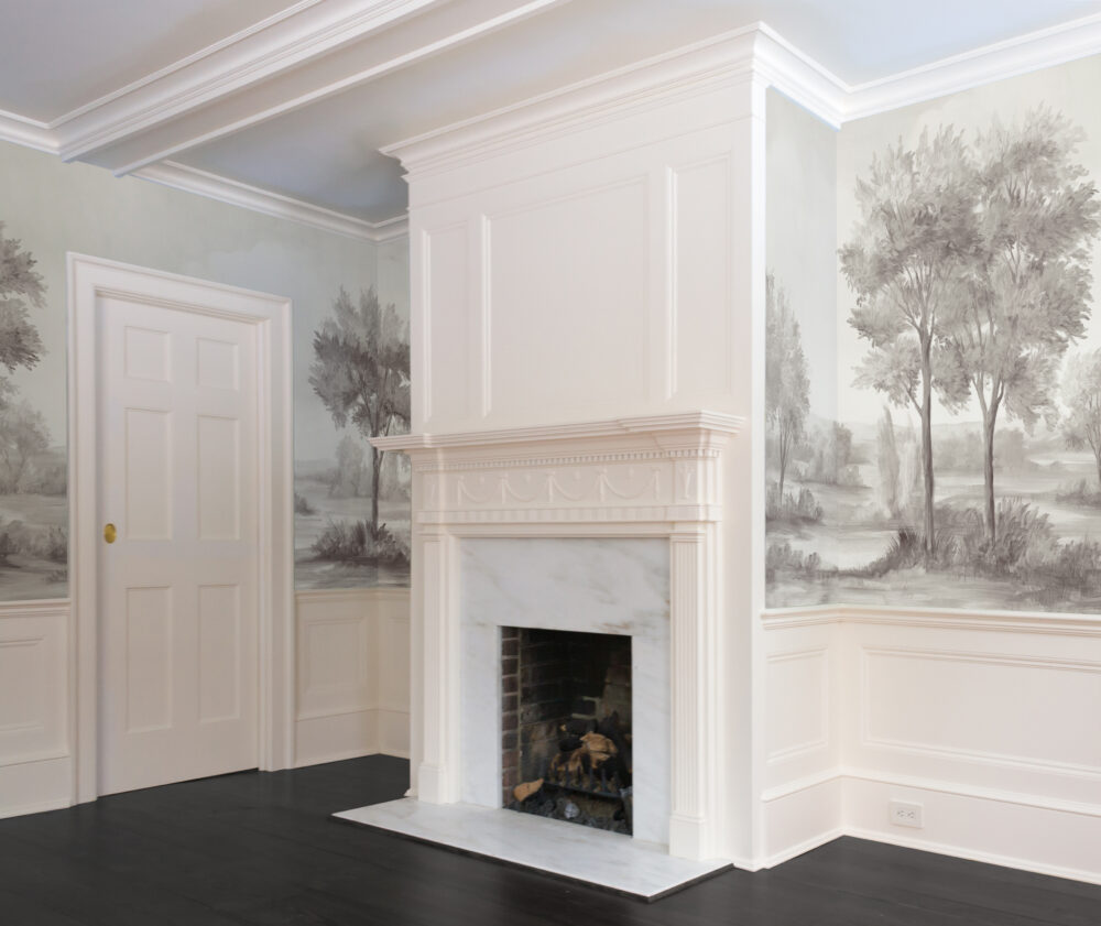 Fireplace in a room with dark floors, eggshell wainscoting and trimming, and a greyscale grisaille painted landscape mural wallpaper
