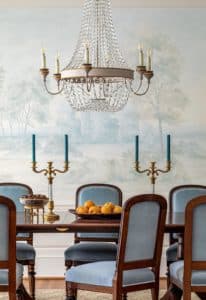 Pale blue velvet chairs around antique dining room table, a crystal chandelier above, with a soft and misty wall mural behind