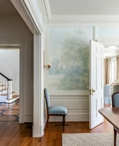 Pale blue velvet antique chair in the corner of a dining room in front of a soft painted mural landscape wallpaper.